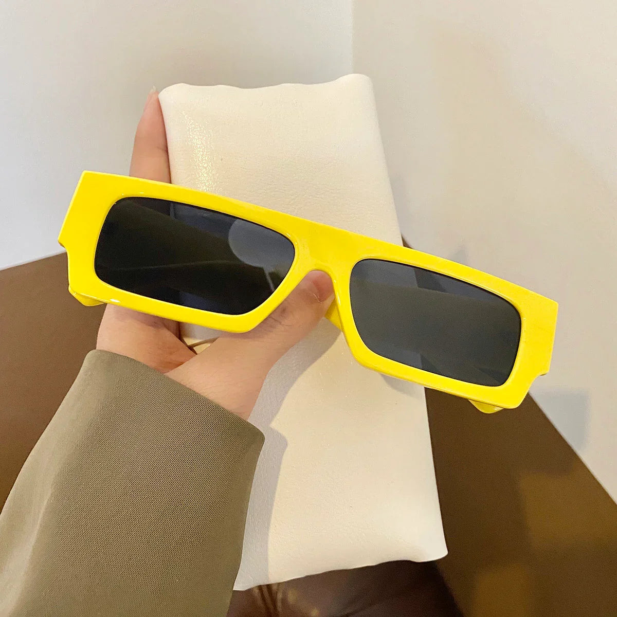 Hand holding bold yellow rectangular sunglasses with dark lenses over a white cushion