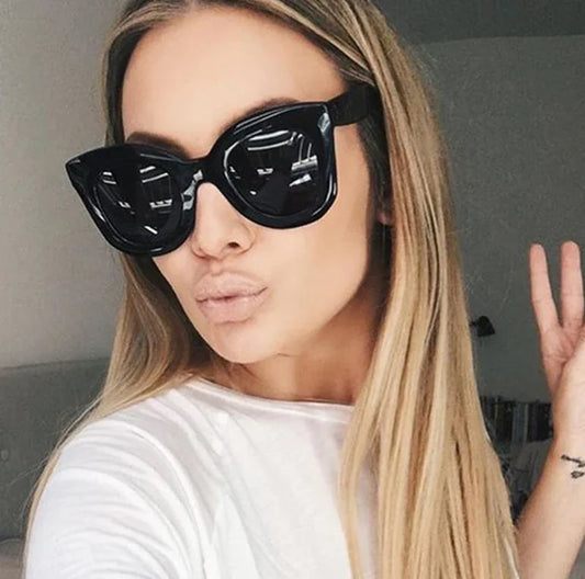 Woman wearing oversized black cat-eye sunglasses and white top posing indoors