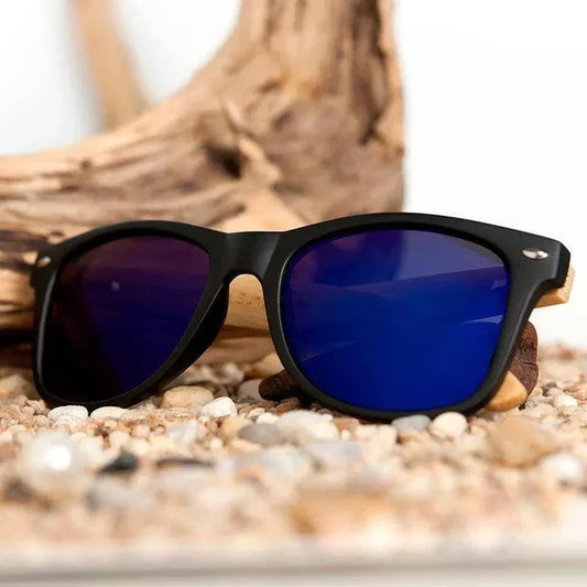 Stylish black sunglasses with blue mirrored lenses on pebbles and driftwood, perfect for sunny days.