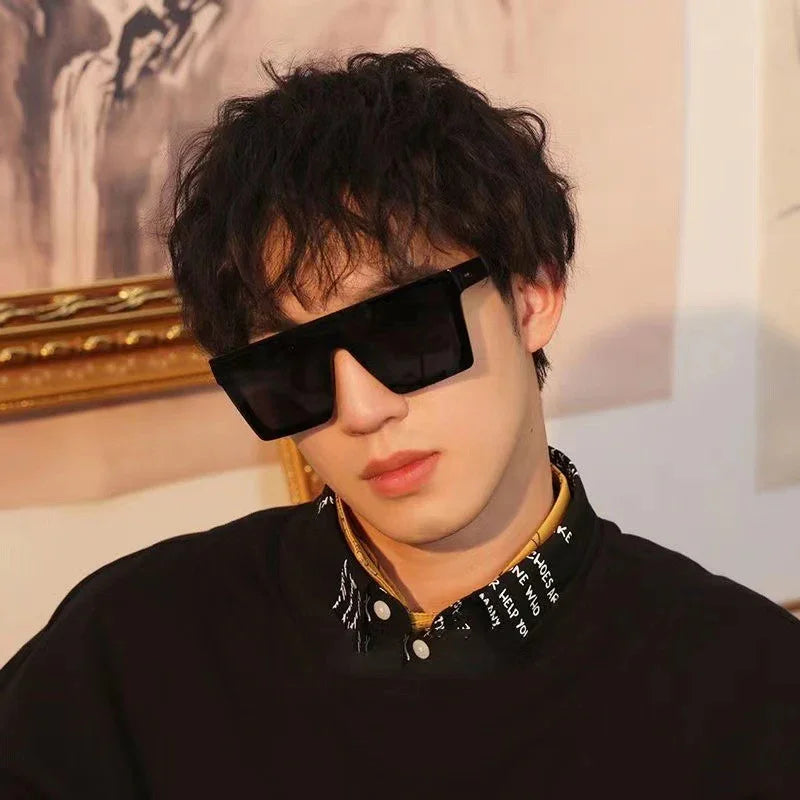 Young man wearing oversized black square sunglasses and a black shirt with patterned collar indoors