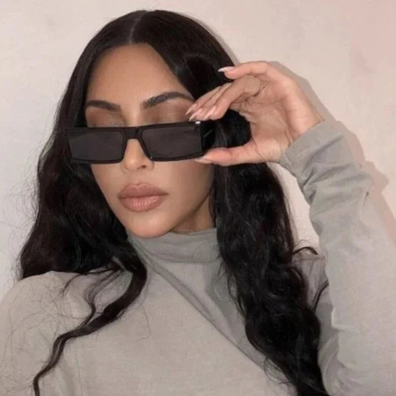 Woman with long dark wavy hair wearing a grey turtleneck and large rectangular black sunglasses