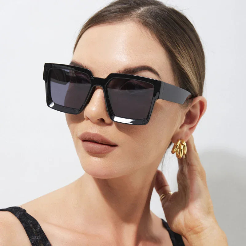 Woman wearing oversized square black sunglasses and gold hoop earrings with hand near ear
