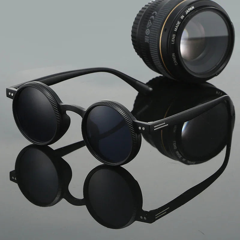Round black sunglasses with textured rims beside a camera lens on reflective surface