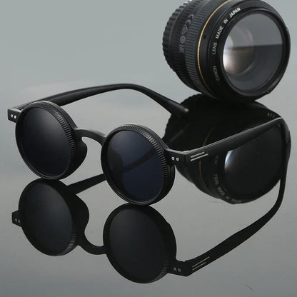 Round black sunglasses with textured rims beside a camera lens on reflective surface