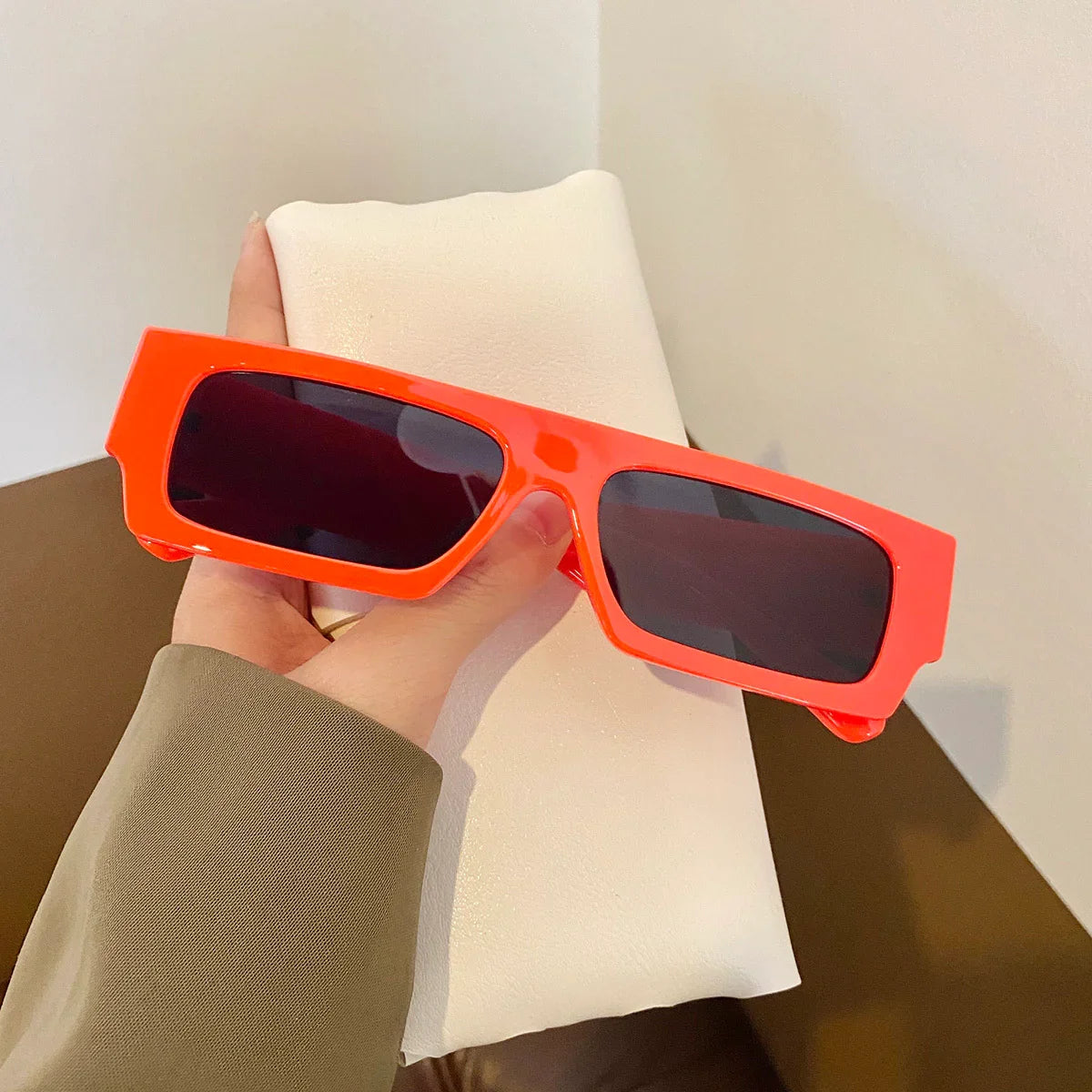 Hand holding bright red rectangular sunglasses with dark lenses against a white cushion background