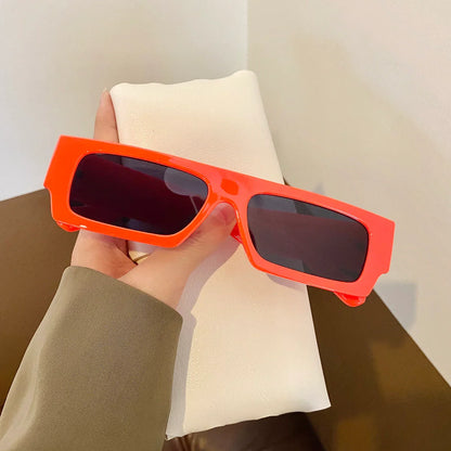 Hand holding bright red rectangular sunglasses with dark lenses against a white cushion background