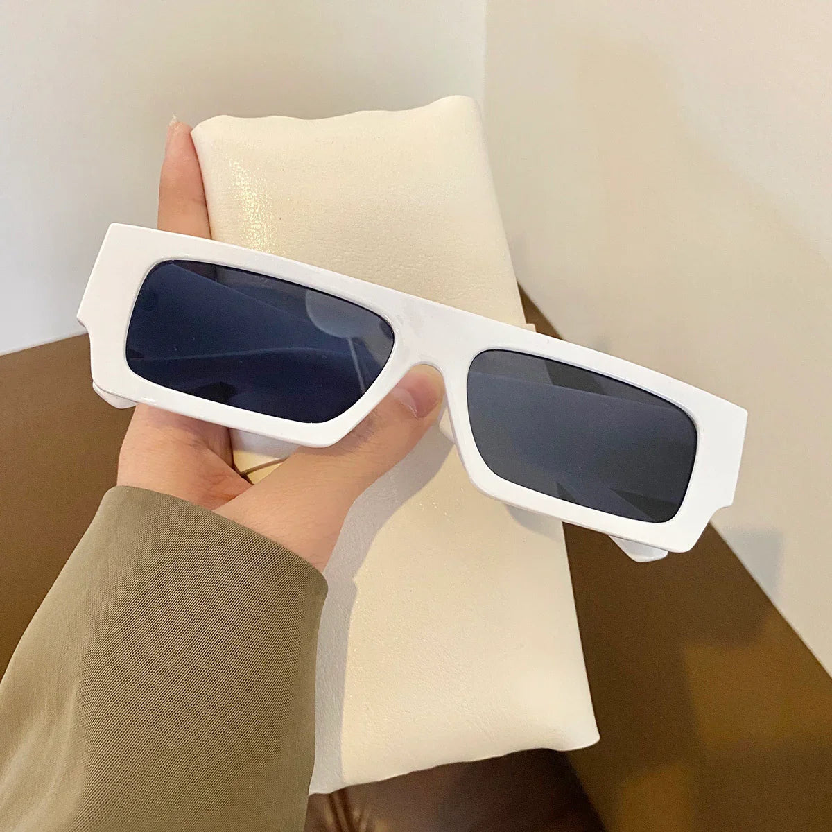 Hand holding white rectangular frame sunglasses with dark lenses over a beige pouch
