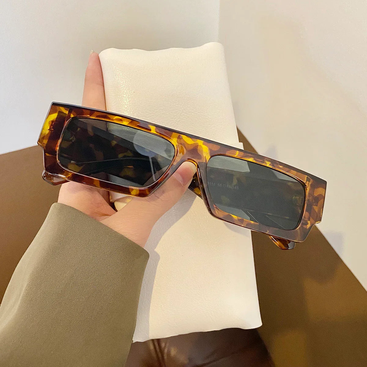 Hand holding rectangular tortoiseshell sunglasses with dark lenses over a white leather surface