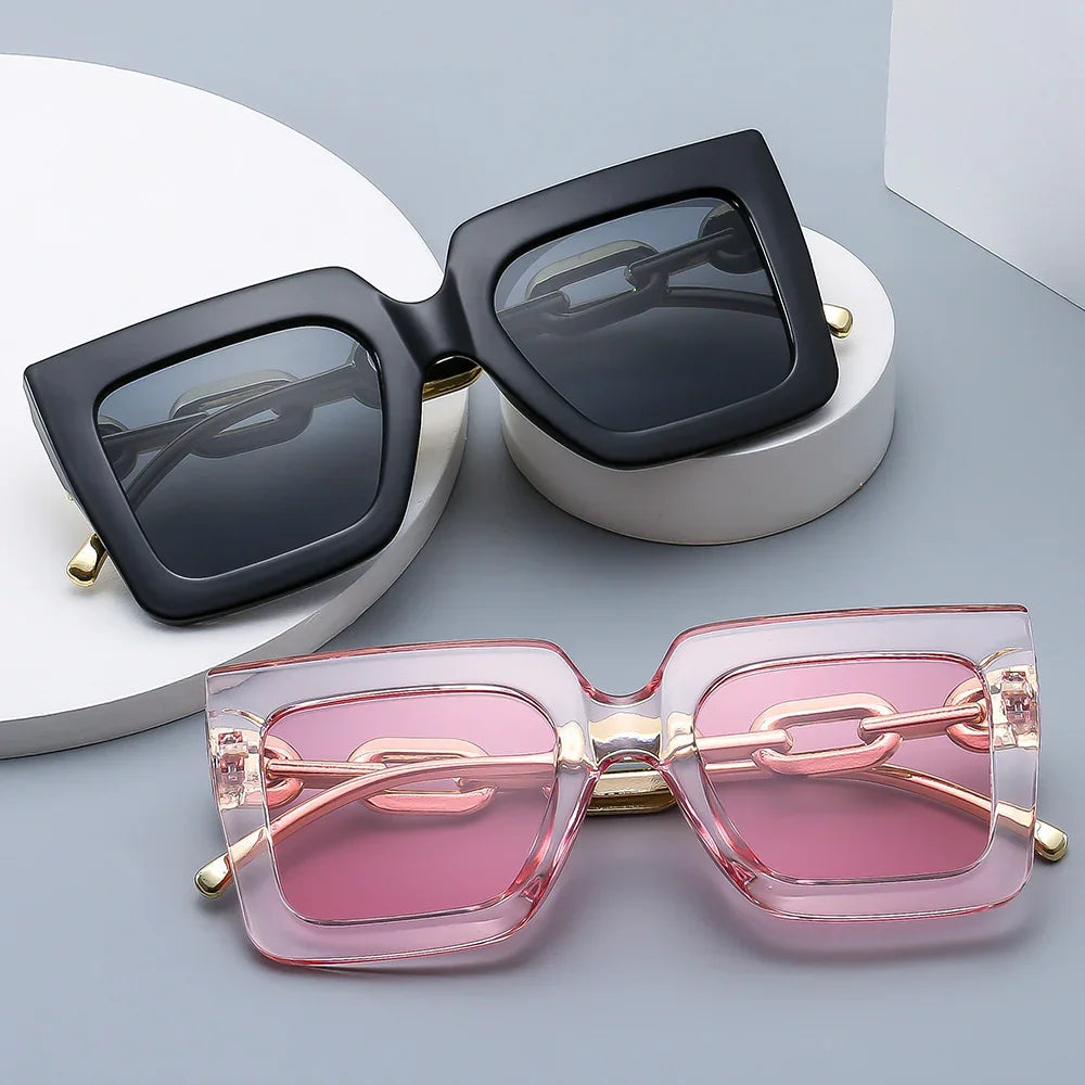 Two pairs of oversized square sunglasses with thick frames; one black with dark lenses, one translucent pink with pink lenses