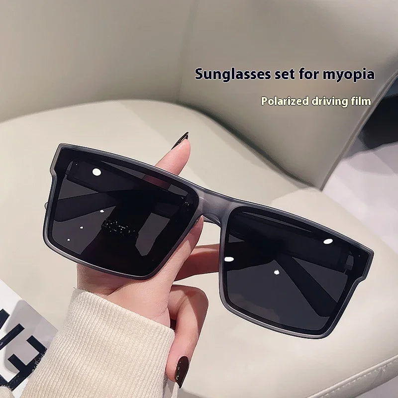 Hand holding square black sunglasses with dark lenses on a beige leather surface