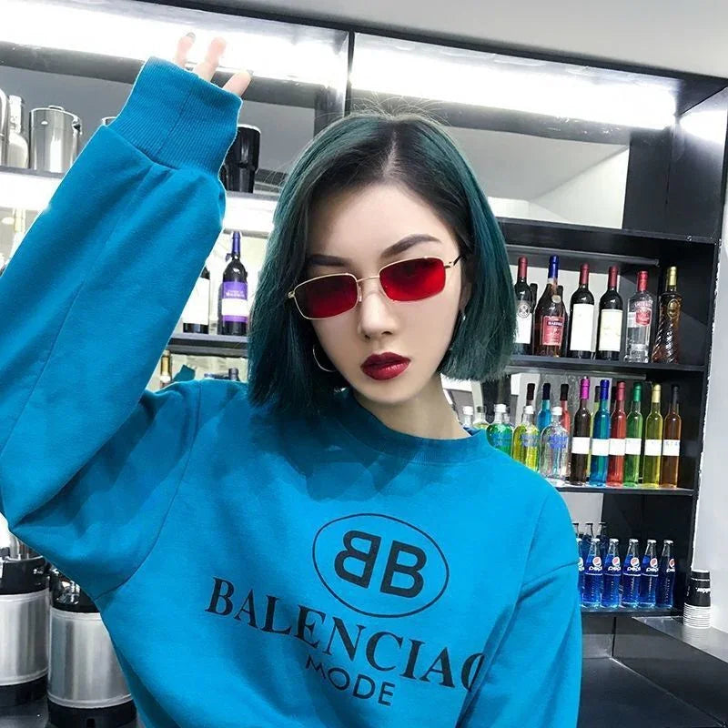 Young woman with blue bob hair wearing red sunglasses and blue sweatshirt in front of liquor bottles