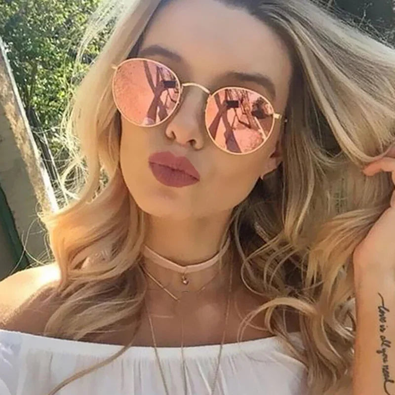 Blonde woman wearing round pink mirrored sunglasses, off-shoulder white top, and layered necklaces outdoors
