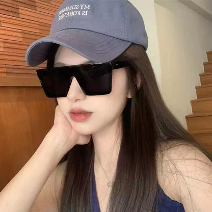 Woman wearing large black sunglasses and a blue baseball cap with long brown hair, indoors near wooden stairs