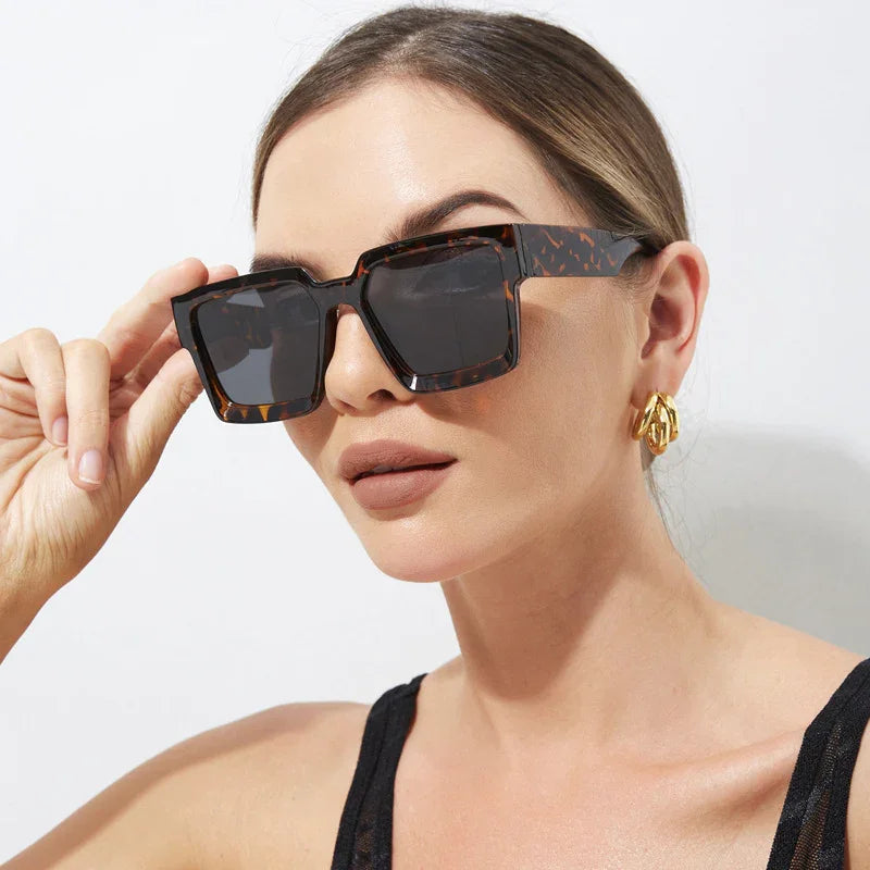 Woman wearing oversized tortoiseshell square sunglasses and gold hoop earrings against white background