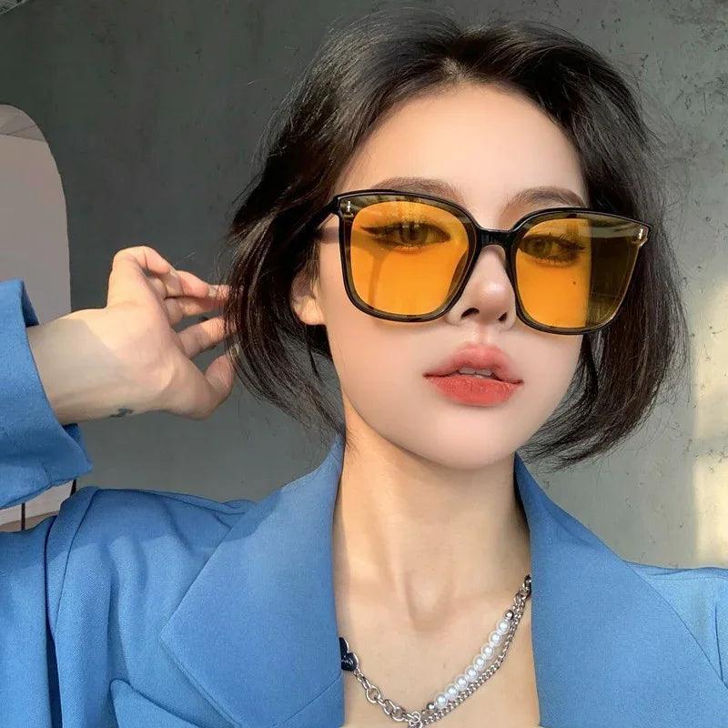Woman wearing stylish oversized yellow-tinted sunglasses with black frames, blue blazer, and pearl necklace