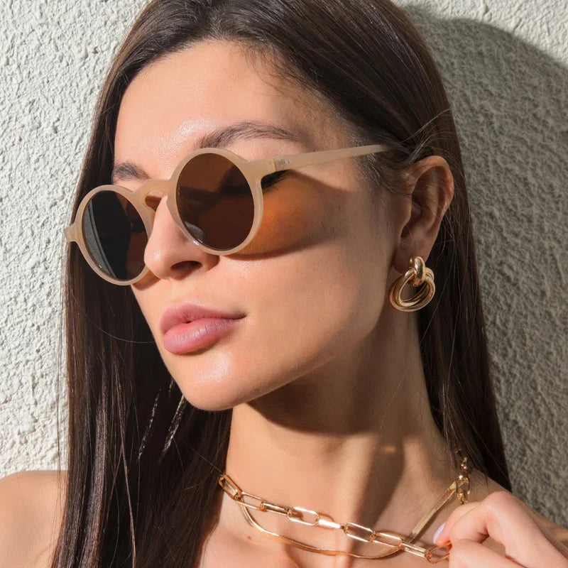 Woman wearing beige round sunglasses and gold hoop earrings with gold chain necklace in natural light