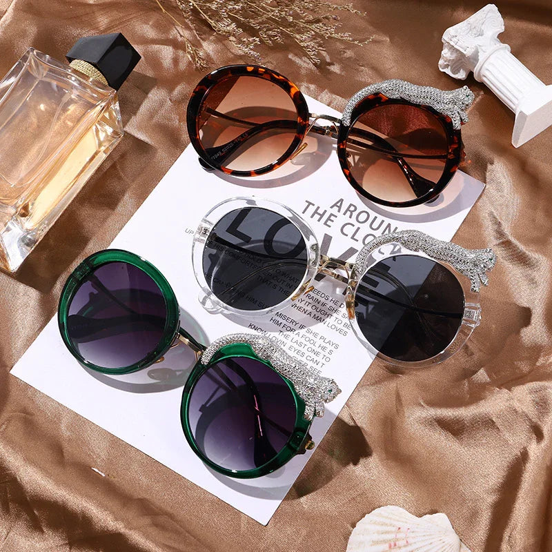 Three pairs of elegant round sunglasses with jeweled animal accents on glossy bronze fabric next to perfume bottle and decor