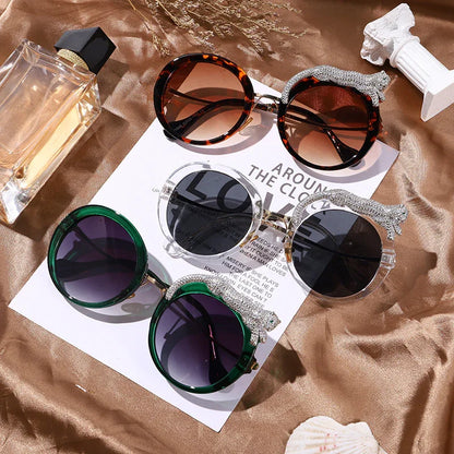 Three pairs of elegant round sunglasses with jeweled animal accents on glossy bronze fabric next to perfume bottle and decor