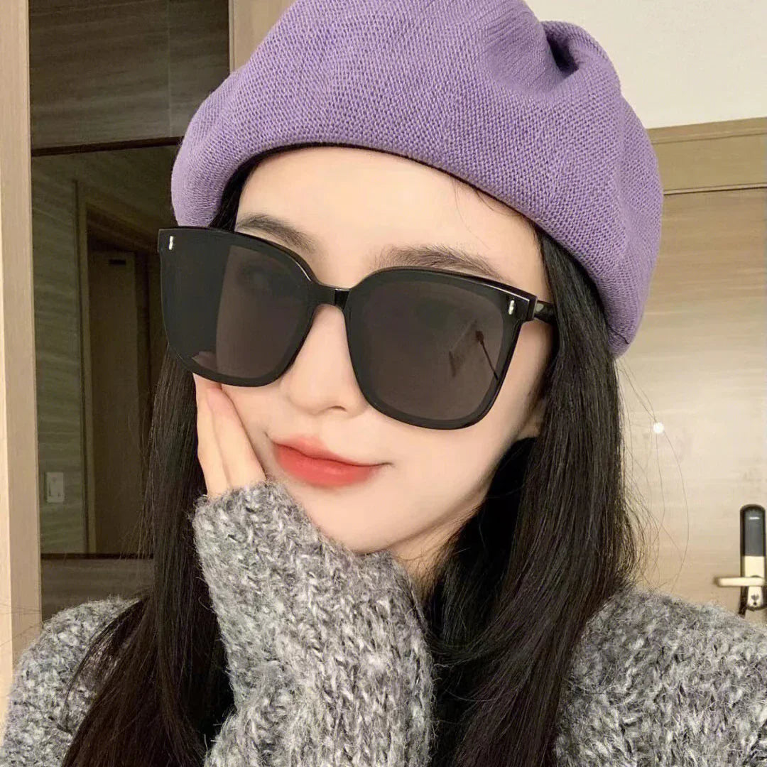 Young woman wearing oversized black sunglasses, purple knit beret, and gray chunky sweater indoors