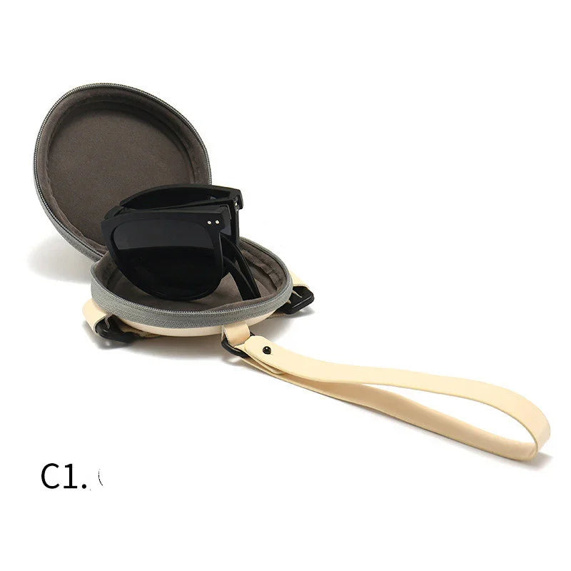 Black sunglasses folded inside a round gray hard case with beige wrist strap on white background
