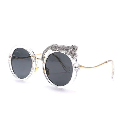 Round sunglasses with clear frames, dark lenses, gold temples, and a decorative silver panther detail