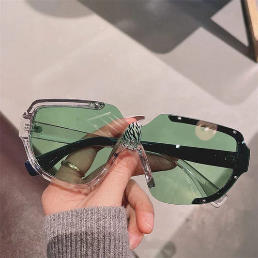 Hand holding large oversized clear frame sunglasses with green tinted lenses and zebra-striped nose piece