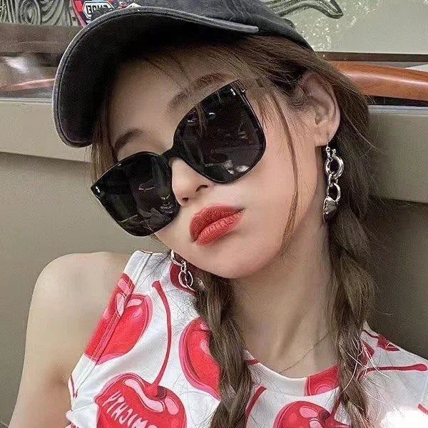 Young woman with braided hair, oversized black sunglasses, red lipstick, cap, cherry print top, and silver hoop earrings