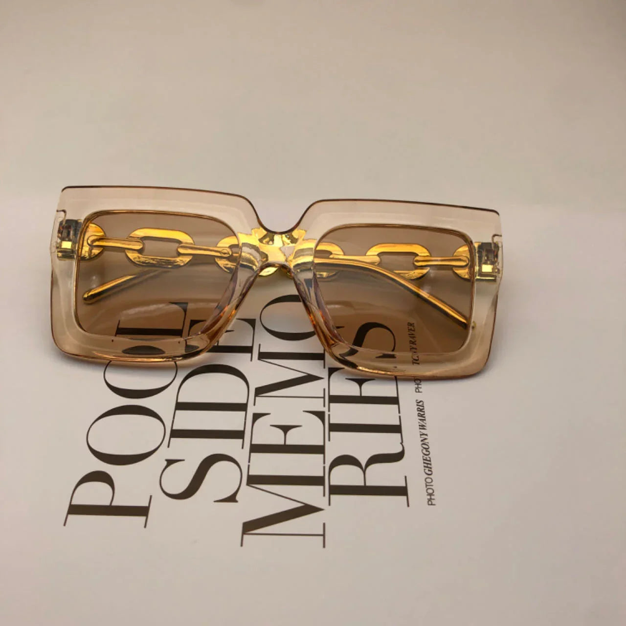 Transparent beige square sunglasses with gold chain detail on temples on a magazine page