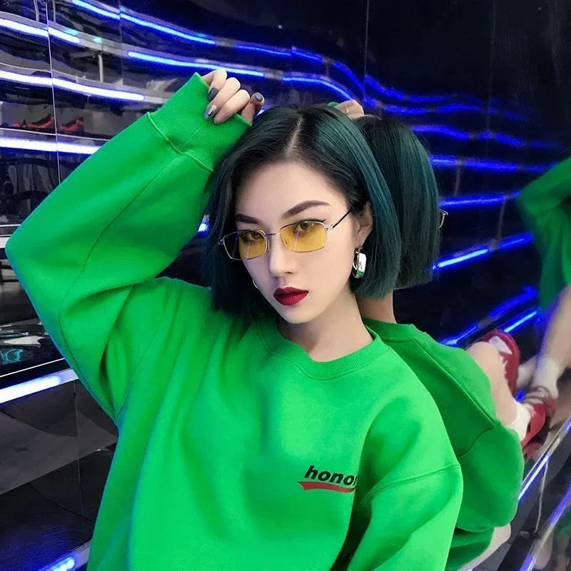 Young woman with short dark hair wearing yellow-tinted glasses and a bright green sweatshirt in neon-lit room