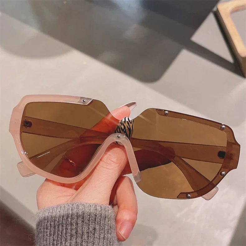 Hand holding oversized brown tinted shield sunglasses with clear geometric frame and metal accents