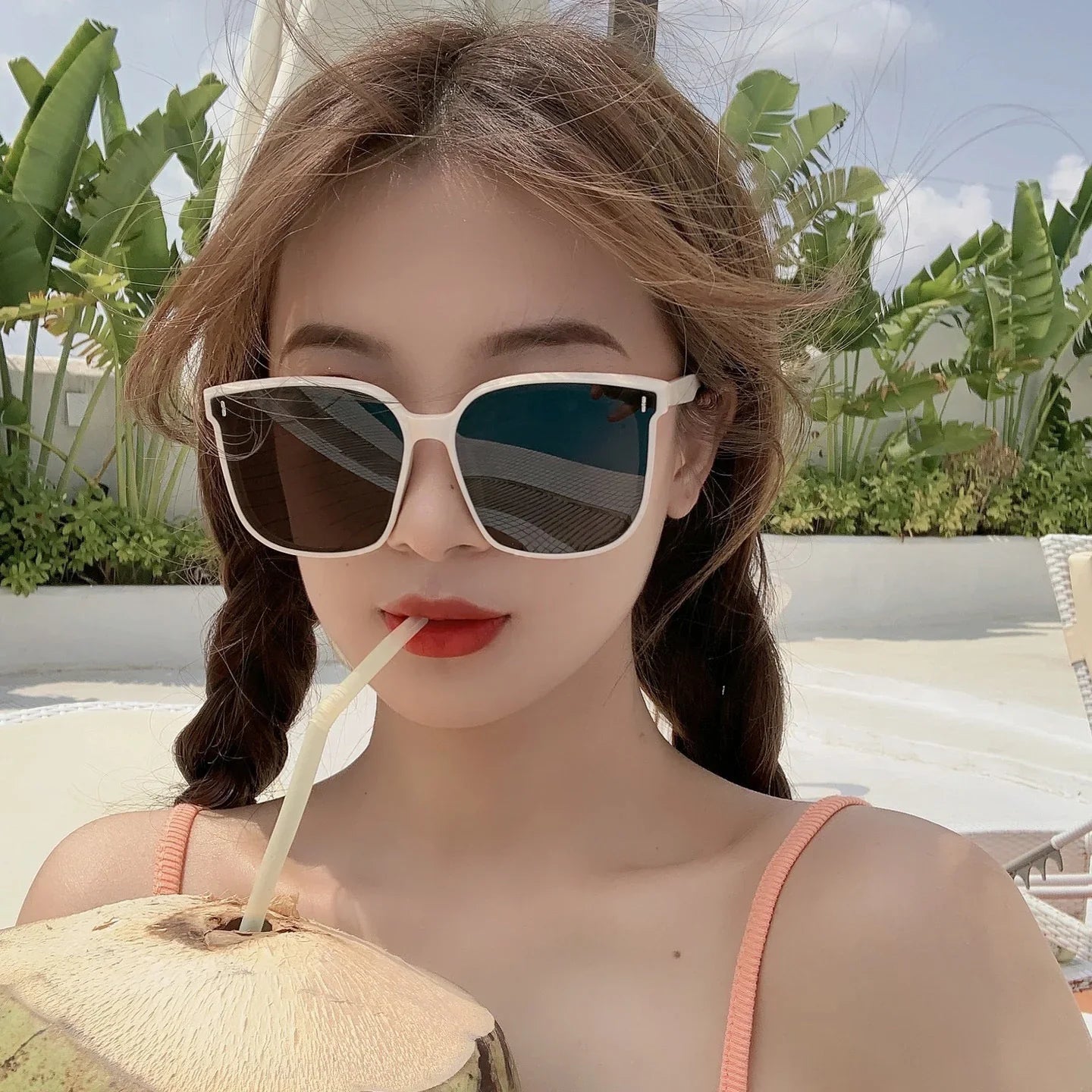 Young woman in pink swimsuit wearing large square sunglasses sipping from a coconut with tropical plants in background