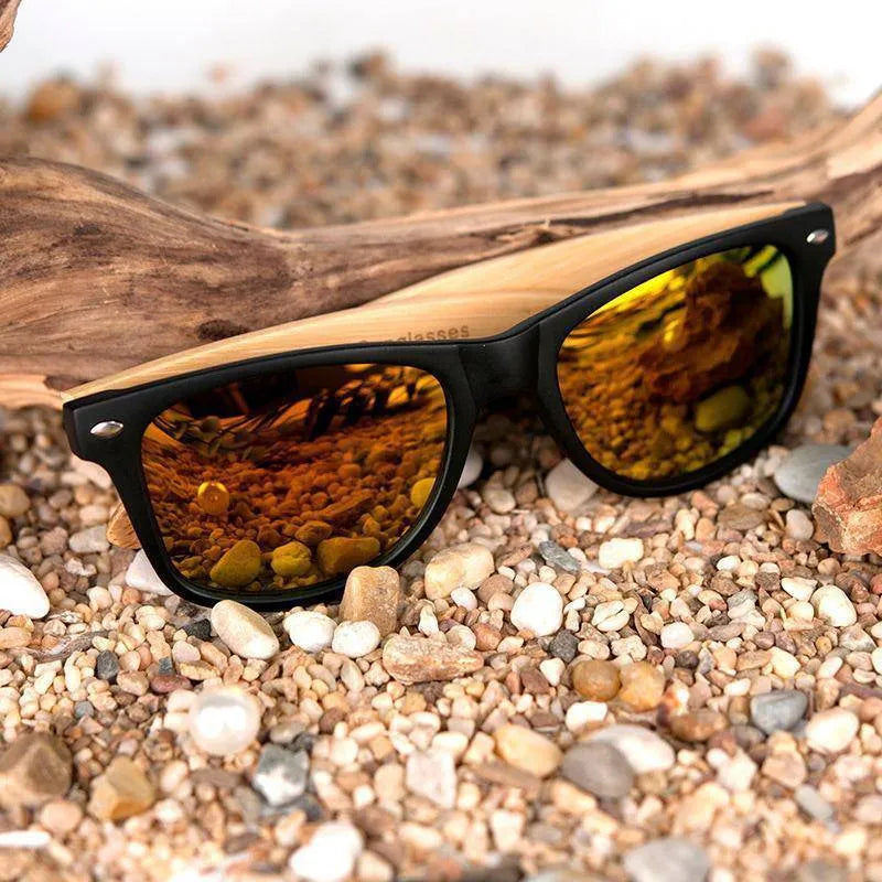 Wood-framed sunglasses with black rims and amber reflective lenses resting on small pebbles and driftwood