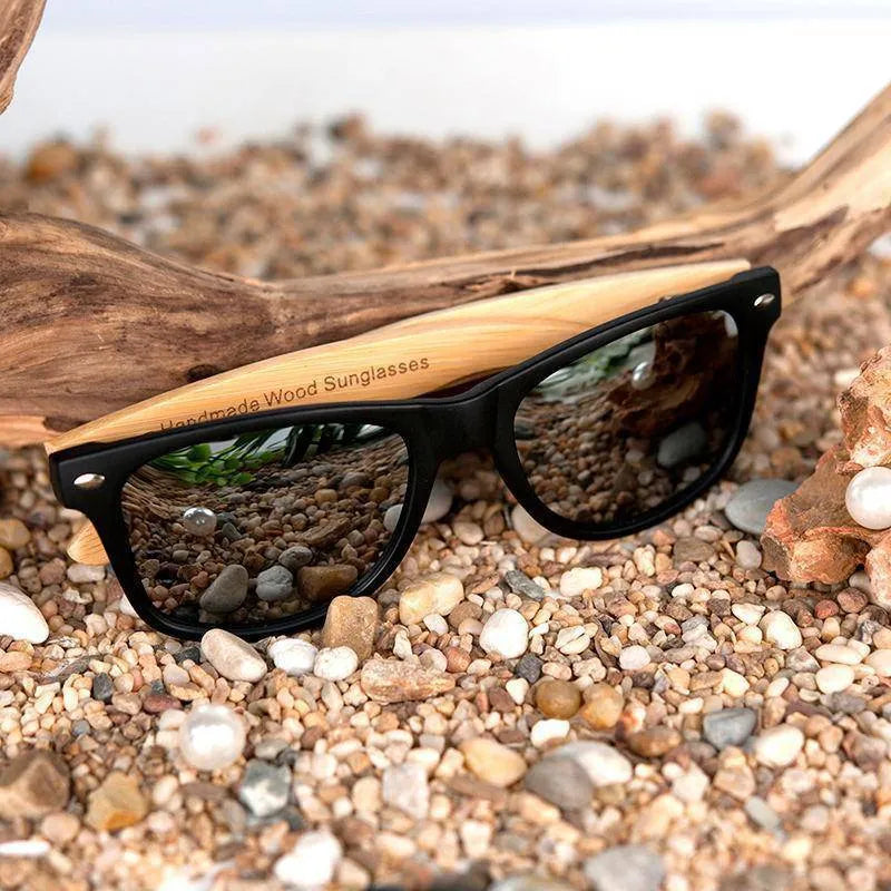 Black frame sunglasses with wooden arms on pebbled beach with driftwood and seashells