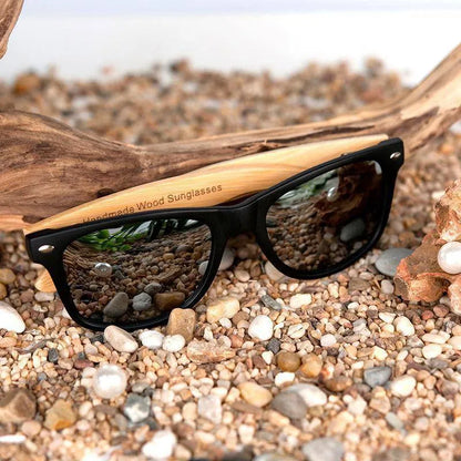 Black frame sunglasses with wooden arms on pebbled beach with driftwood and seashells