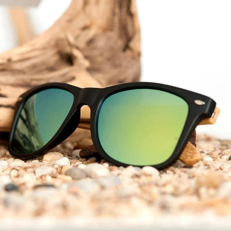 Black frame sunglasses with green reflective lenses resting on pebbles with driftwood background