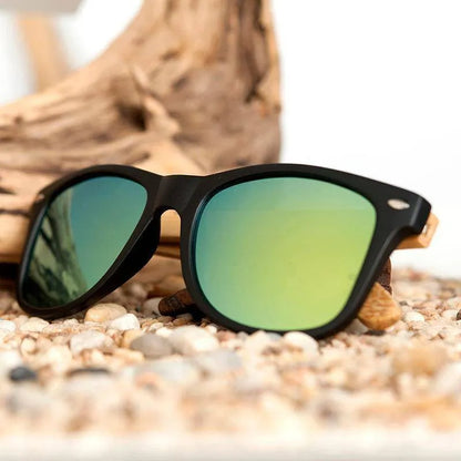 Black frame sunglasses with green reflective lenses resting on pebbles with driftwood background